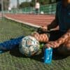 A soccer player takes a break, sitting on a grassy field with a soccer ball and water bottle.