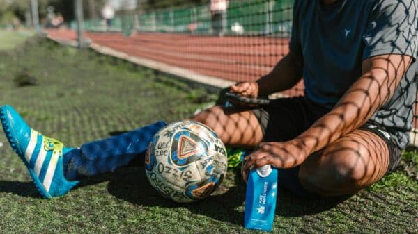 A soccer player takes a break, sitting on a grassy field with a soccer ball and water bottle.